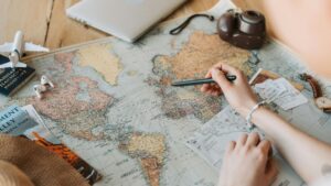 世界地図の上でペンを持ち、旅行ルートを指し示す手。周囲にはパスポート、搭乗券、カメラ、時計、イヤホン、ミニチュア飛行機が置かれている。