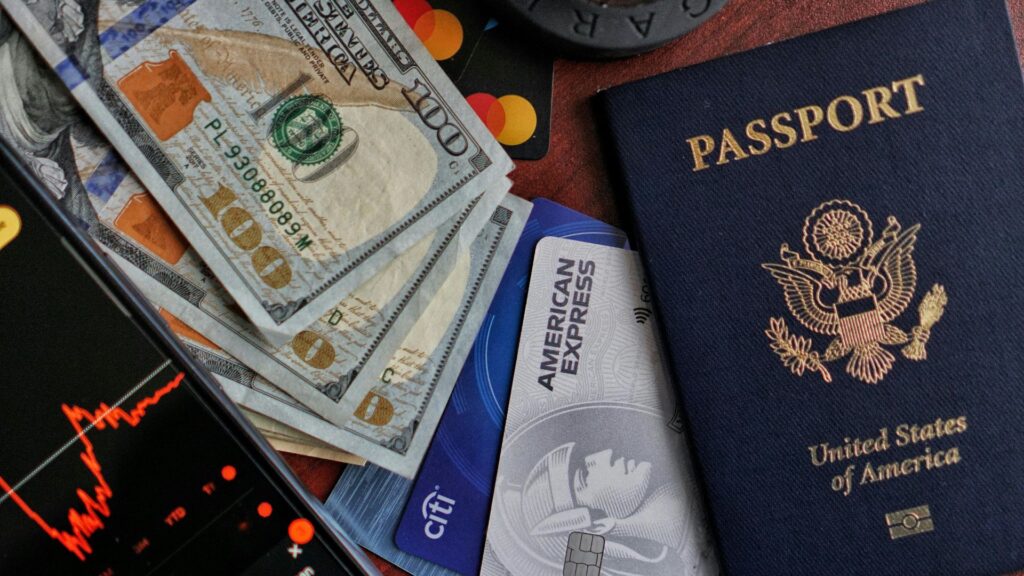 米ドル紙幣、American ExpressとCitiのクレジットカード、マスターカードのロゴ、米国パスポート、株価チャートを表示したスマホが机上に並ぶ