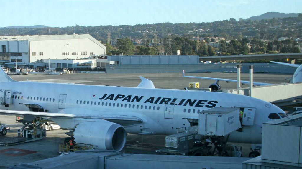空港のゲートに駐機する日本航空（JAL）のボーイング787。機体側面に「JAPAN AIRLINES」の文字、手前に貨物の積み込み車両